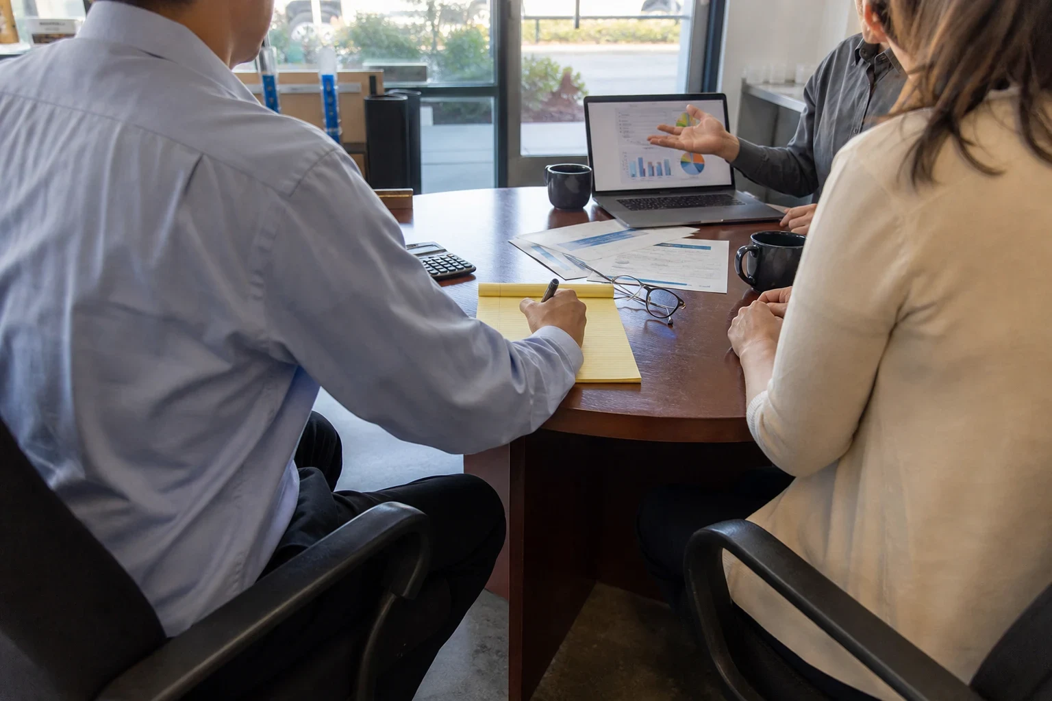 Man in a light blue button-down takes notes on a yellow legal pad at a round office table while a woman listens and another person points to charts on a laptop.